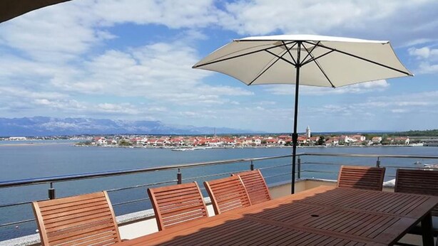 Una terraza de madera con una mesa, sillas y una sombrilla, con vista a un cuerpo de agua y edificios al fondo.