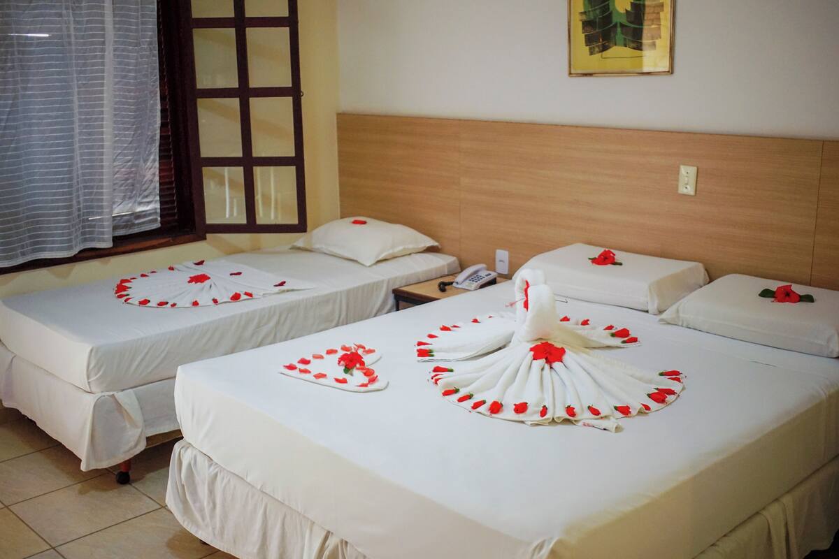 A hotel room with two single beds, each with a white bedspread and red rose decorations. There is a wooden headboard, a bedside table with a phone, and a window with a curtain.