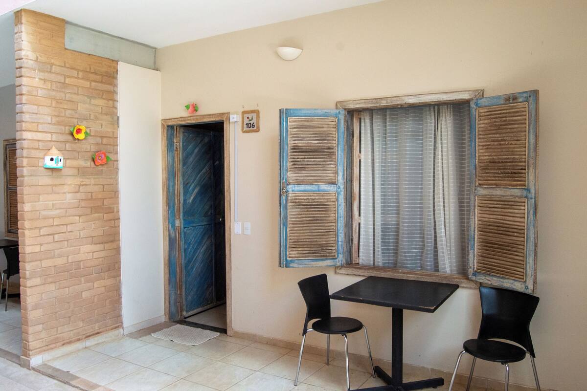 A room with a brick wall, a door, two windows with shutters, a small table, and two chairs.