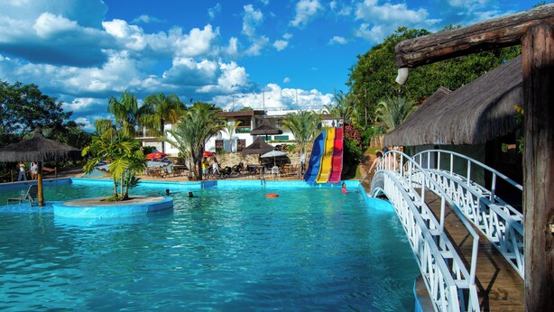 A swimming pool with a wooden bridge, palm trees, and a thatched-roof building. - Vila Velluti Hotel SPA (Gama)