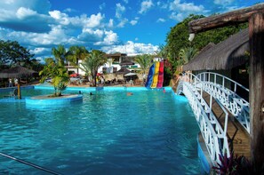 Una piscina con un puente de madera, palmeras y un edificio con techo de paja.