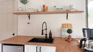 A kitchen with a wooden countertop, a black sink, and a white cabinet. There are two wooden shelves above the sink, holding glassware and a potted plant. A black wall-mounted light fixture is above the shelves. - The Hikers Haven  (Thompson Falls)