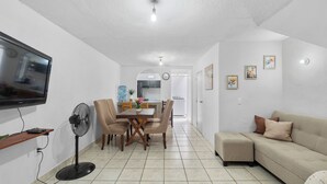 Un salon moderne avec un téléviseur à écran plat, un ventilateur sur pied, un coin repas avec une table et des chaises, un canapé et une cuisine en arrière-plan.