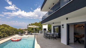 Un área de piscina moderna al aire libre con vista despejada a la ciudad, un comedor con mesas exteriores y un amplio ventanal con vistas a la piscina.