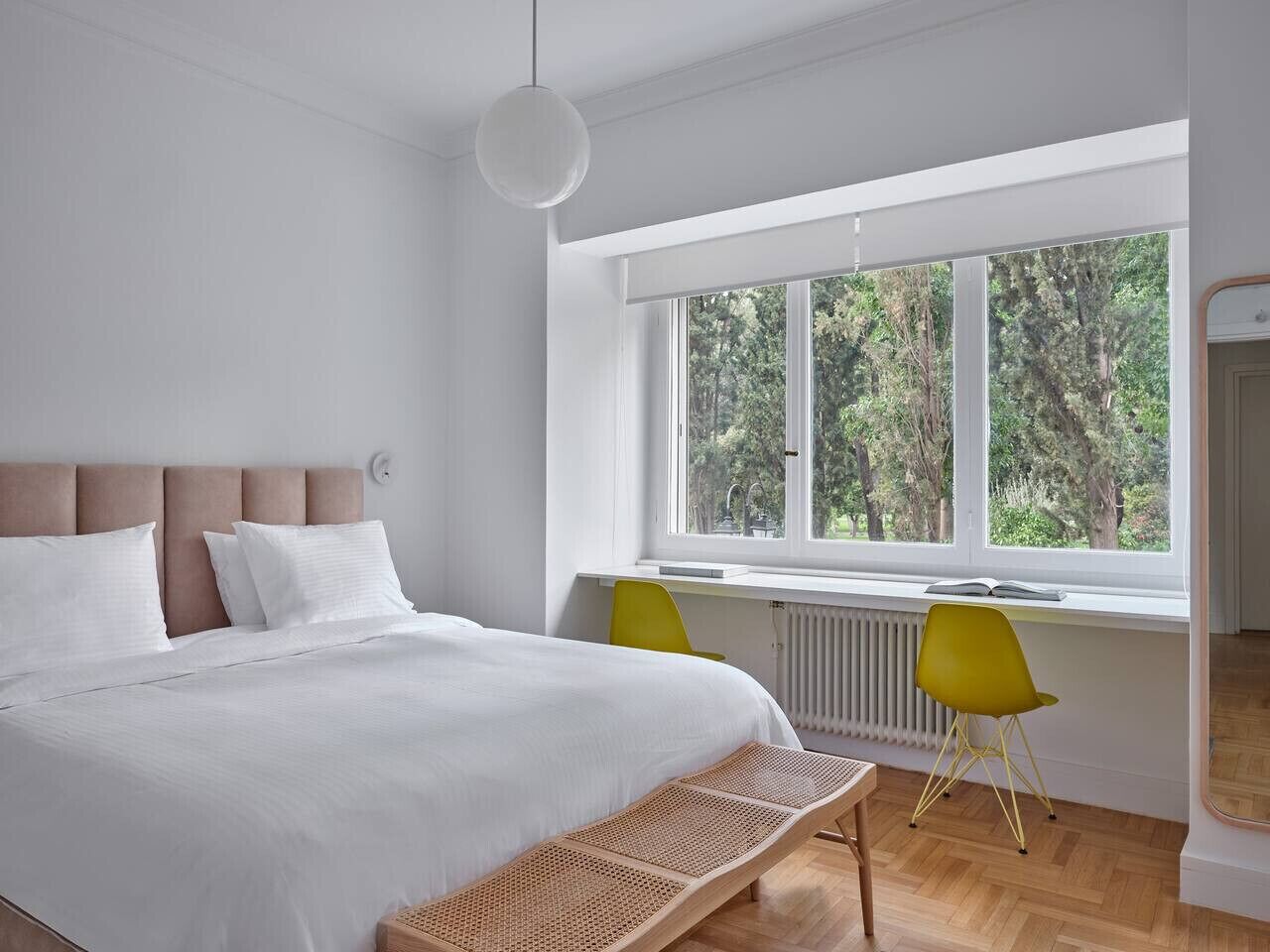 A modern bedroom with a large bed, a window with a view of trees, a bright yellow chair, and a white radiator.
