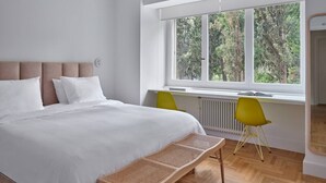 A modern bedroom with a large bed, a window with a view of trees, a bright yellow chair, and a white radiator. - Anaktora Gardens Apt (Athina)