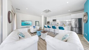 Una sala de estar moderna con sofás blancos, una mesa de centro y un televisor que muestra un barco en la pantalla. Se ve una cocina con gabinetes blancos y un refrigerador de acero inoxidable al fondo.