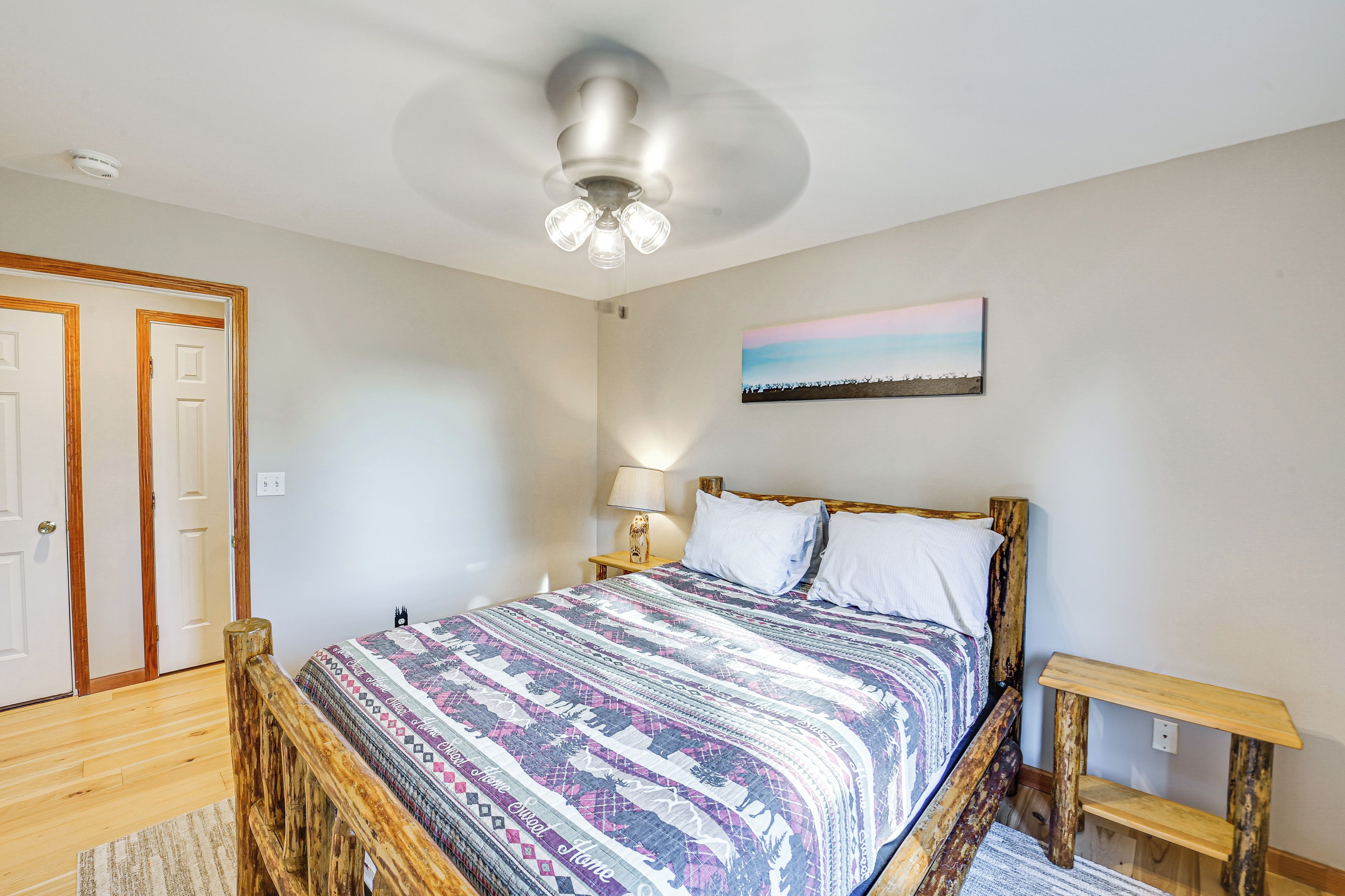 Un dormitorio con cama de madera, ventilador de techo, mesita de noche, lámpara y un cuadro en la pared.