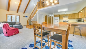Een ruime woonkamer met een hoge plafond, houten balken, een eettafel met stoelen, een rode fauteuil en een keuken met houten kastjes en apparatuur.