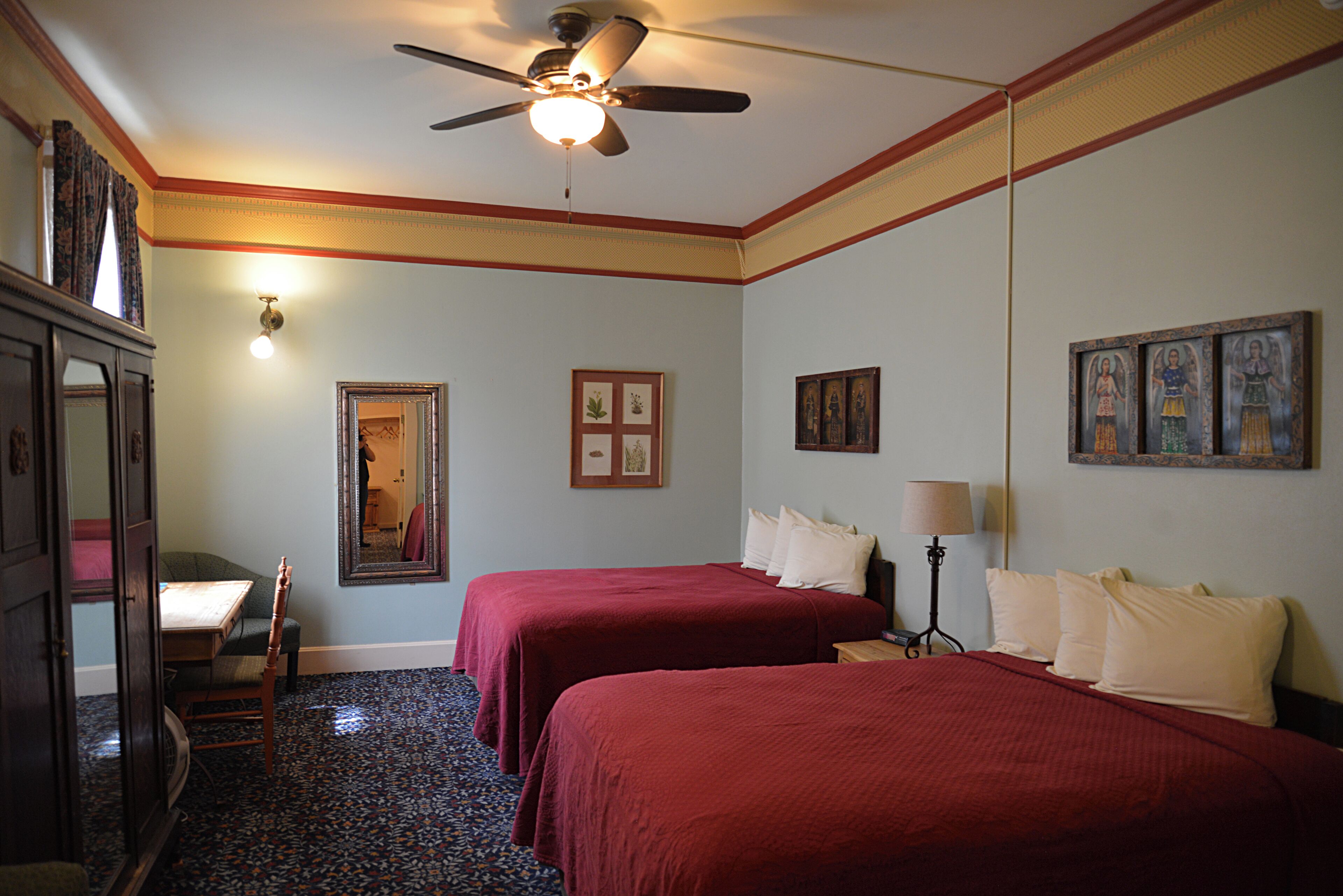 A hotel room with two beds, a ceiling fan, a desk with a chair, a dresser, and framed pictures on the walls.