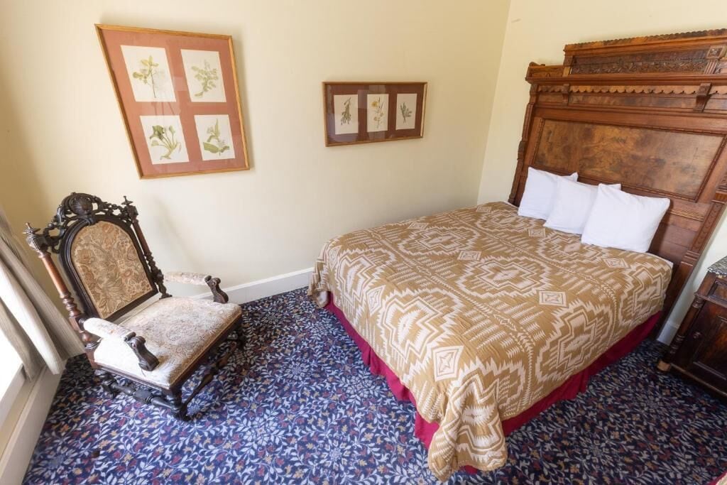 A bedroom with a patterned carpet, a wooden bed with white pillows, a wooden chair, and framed botanical prints on the wall.