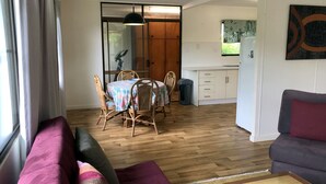 A modern living room with a sofa, a dining table with chairs, a kitchen area, and a sliding glass door leading to a balcony. - Space and tranquillity at Red Door Cottage (Cooktown)