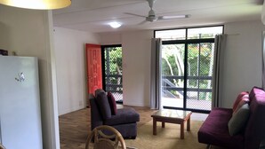 A living room with a wooden floor, a dining table with a floral tablecloth, two sofas, a ceiling fan, and a large window with a view of greenery. - Space and tranquillity at Red Door Cottage (Cooktown)