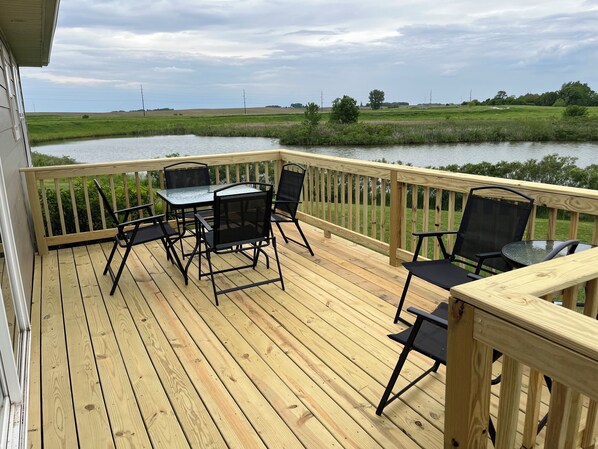 Une terrasse en bois avec une table et des chaises, donnant sur une étendue d’eau et des champs.