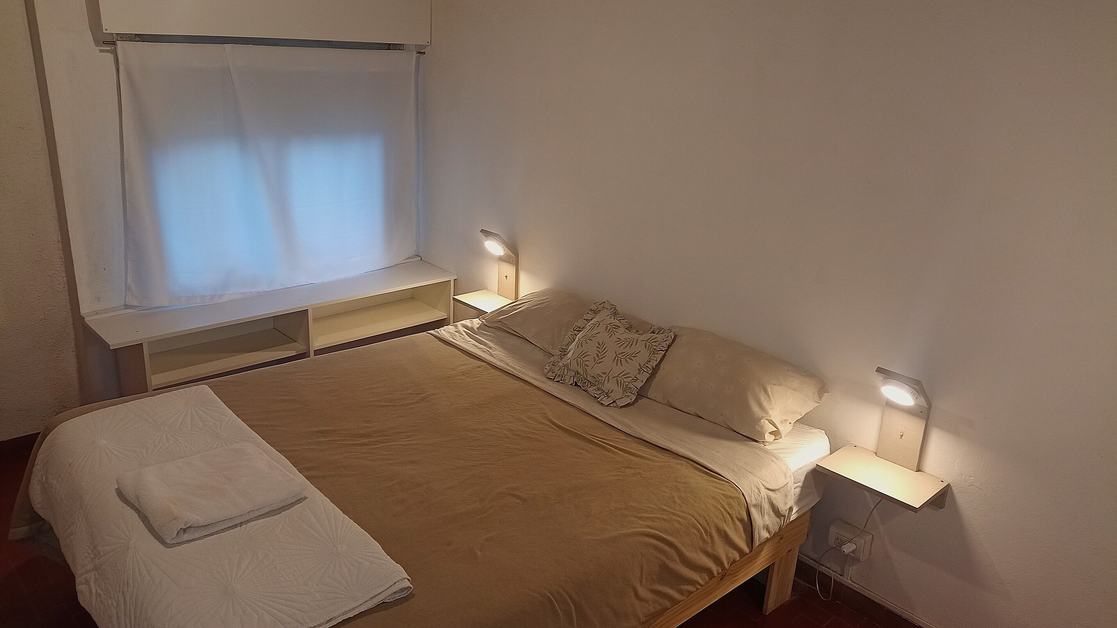 Un lit bien fait, avec une couverture et des oreillers, des lampes de chevet, une fenêtre avec un rideau et une petite étagère sous la fenêtre.
