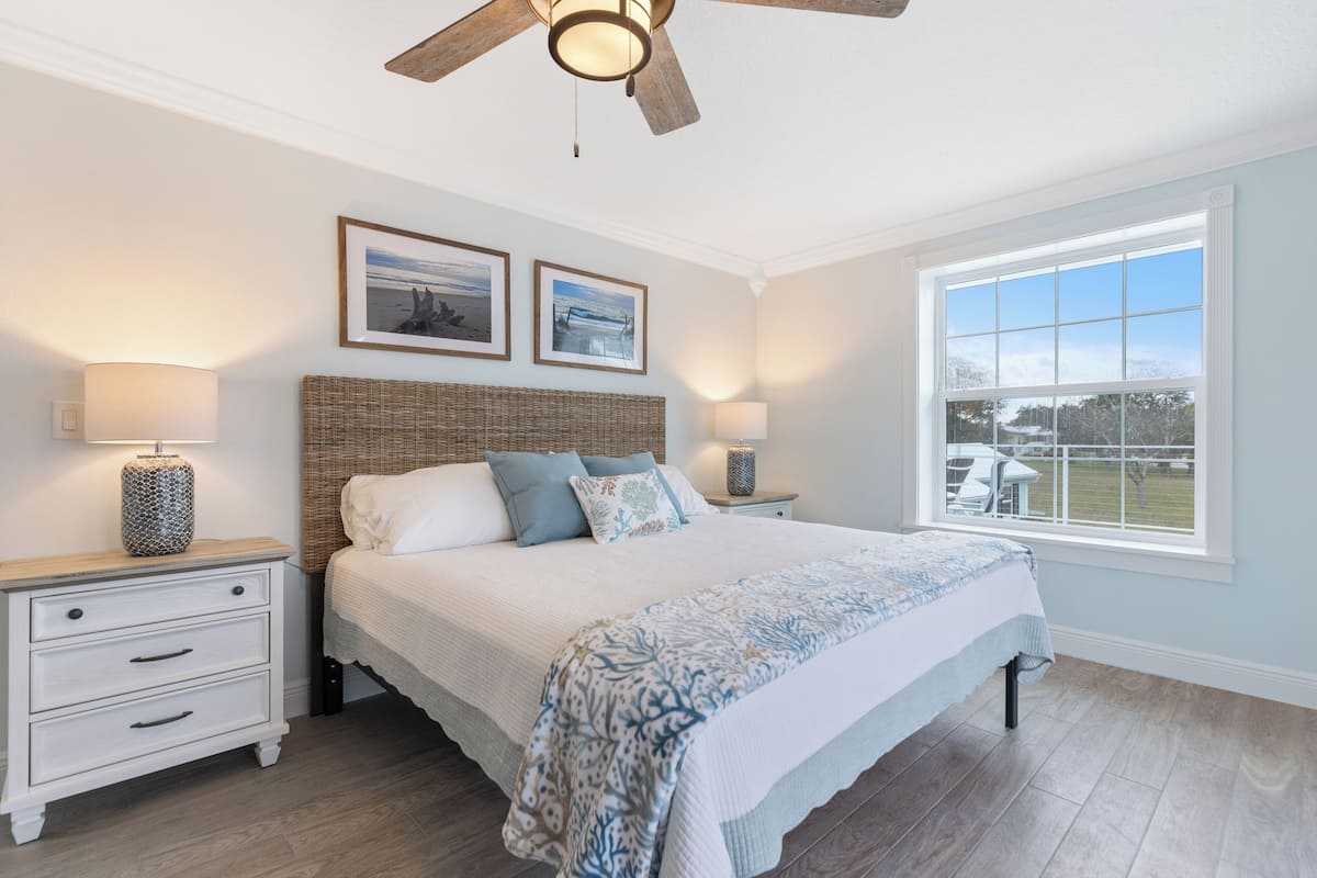 A bedroom with a large bed, two bedside tables, a ceiling fan, and a window with a view of the outdoors.