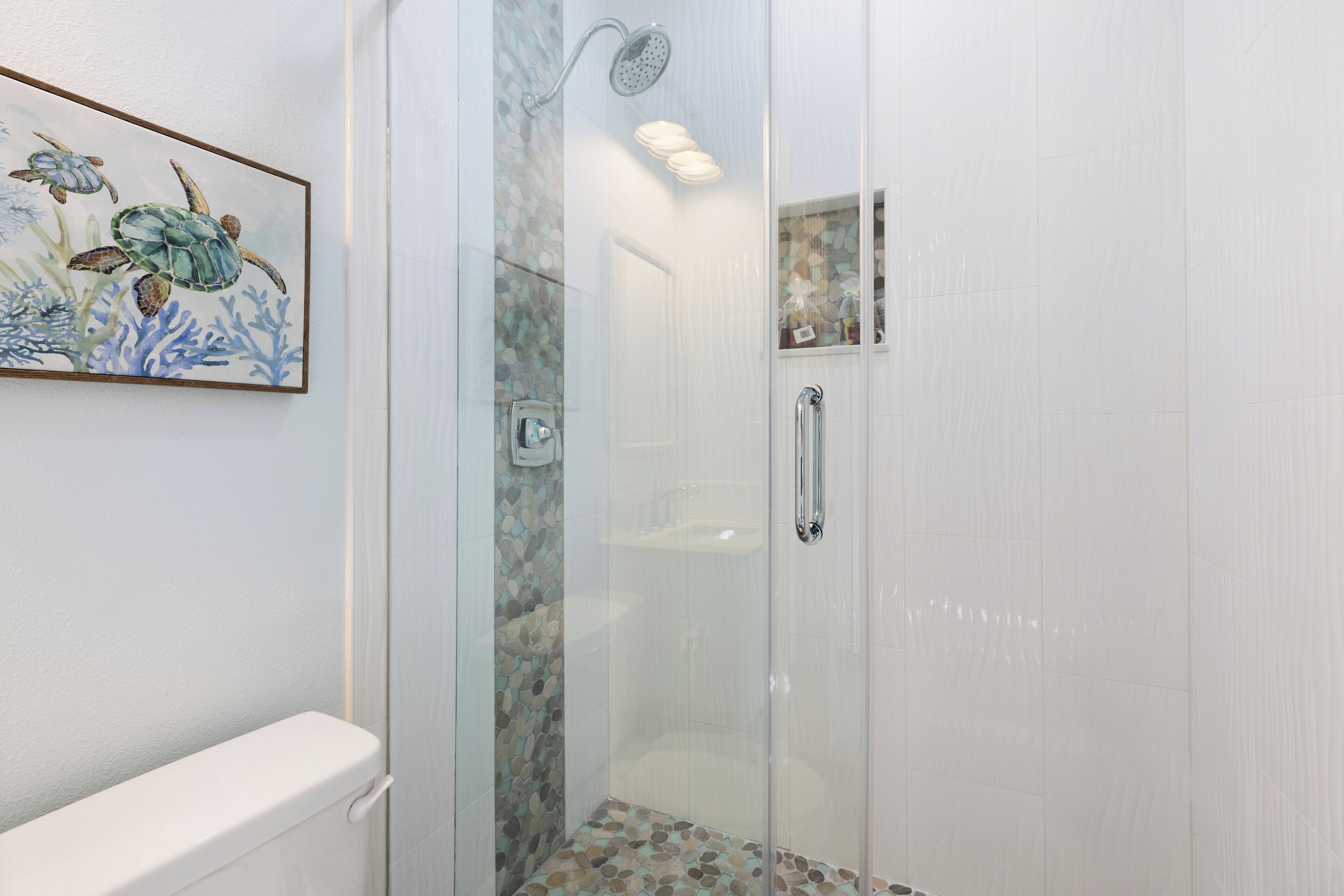A glass-enclosed shower with a tiled floor, a white toilet, and a framed seascape painting on the wall.