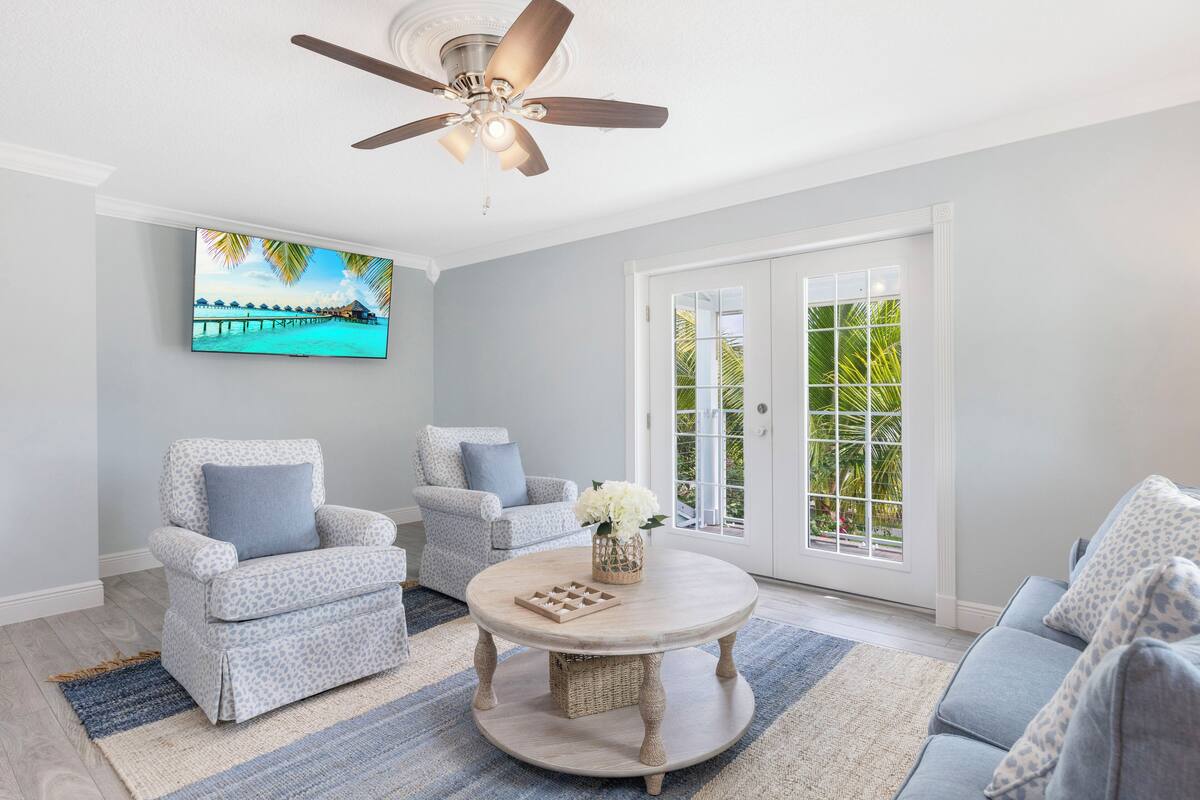 A living room with a ceiling fan, two armchairs, a round coffee table, and a large window with a view of palm trees.