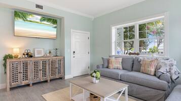 Una sala de estar con un sofá gris, una mesa de centro, un televisor montado en la pared y una ventana con vistas a árboles y una casa.