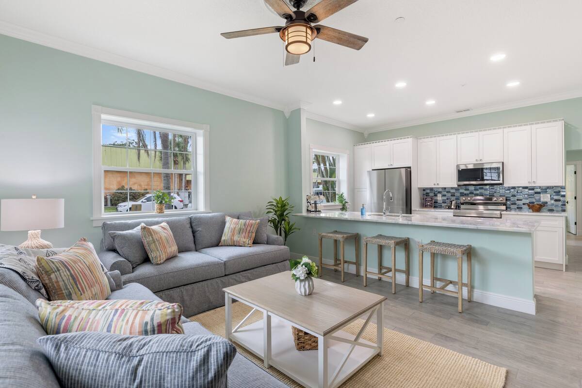 A modern living room with a grey sofa, a coffee table, and a kitchen area with white cabinets and a large island.