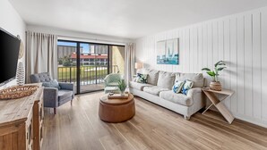 Un salon moderne avec un canapé, une table basse et un meuble pour la télévision. Il y a une grande fenêtre avec des rideaux, un tableau au mur et une plante sur une table d’appoint.