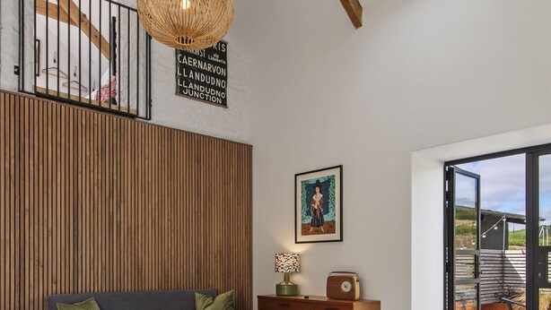 A spacious living room with a high ceiling, wooden beams, a large window, a sofa, a coffee table, and a wicker pendant light. - Cartws at Pentrecelyn- A Stylish Countryside Haven (Carmarthen)