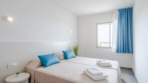 A hotel room with two beds, a small round table, and a window with blue curtains. - APCOSTAS Blue Beach two bedrooms (Salou)