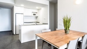 A modern kitchen with white cabinets, a built-in microwave, and a sink. There is a wooden dining table with chairs and a potted plant. - APCOSTAS Blue Beach two bedrooms (Salou)