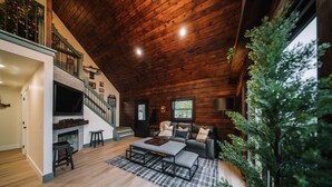 Un salon spacieux avec un haut plafond, des murs en bois, une cheminée, un canapé et un téléviseur.