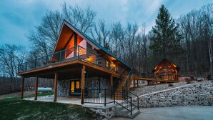 Une cabane en rondins dotée d’un balcon et d’une structure en bois séparée, le tout entouré d’un mur de pierres et d’une zone boisée.
