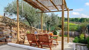 An outdoor dining area with a stone wall, wooden chairs, and a table set with a bottle, glasses, and fruits. - Casa do Jardim Secreto - Monsaraz (Corval)