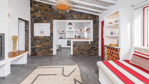 A modern living room with a stone accent wall, a kitchen in the background, and a red and white striped sofa. - Casa do Jardim Secreto - Monsaraz (Corval)