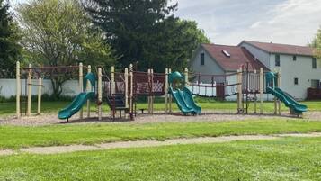 A playground with slides and climbing structures, surrounded by a grassy area and a building in the background.