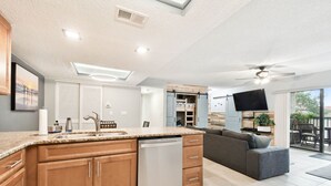 Eine moderne Küche mit Holzschränken, einem Geschirrspüler und einer großen Kücheninsel. Im Wohnbereich befindet sich eine graue Couch und ein Deckenventilator.