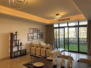 A modern living room with a sofa set, a dining table with plates and vases, a bookshelf, and framed pictures on the wall. - Access - lux smart home with garden at cfc 2bedr (Mivida New Cairo)