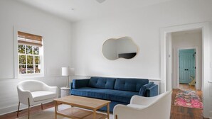 Una sala de estar moderna con un sofá azul, sillones blancos, una mesa de centro de madera y un espejo de pared original.