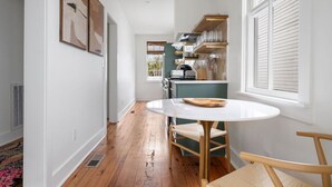 Un pasillo estrecho con una mesa redonda y sillas, una zona de cocina con estantes de madera, y un ventanal con contraventanas.