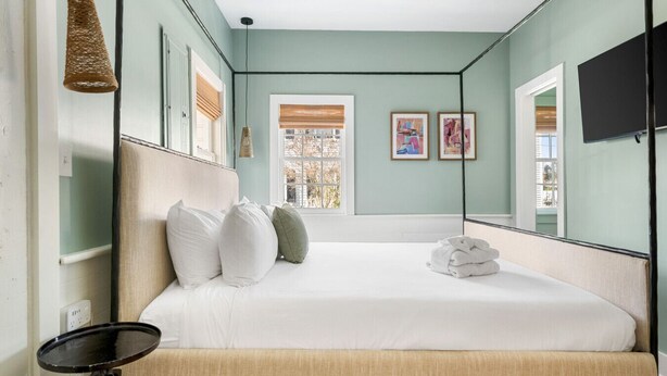 Un dormitorio con cama, almohadas, una mesita de noche, una ventana con persianas y un televisor montado en la pared.