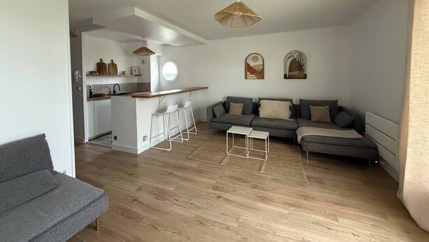 A modern living space with a kitchen, grey sofas, a dining area, and a hanging light fixture.