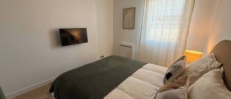 Ein Schlafzimmer mit Bett, Flachbildfernseher an der Wand, einem Fenster mit Vorhängen und einer Nachttischlampe.