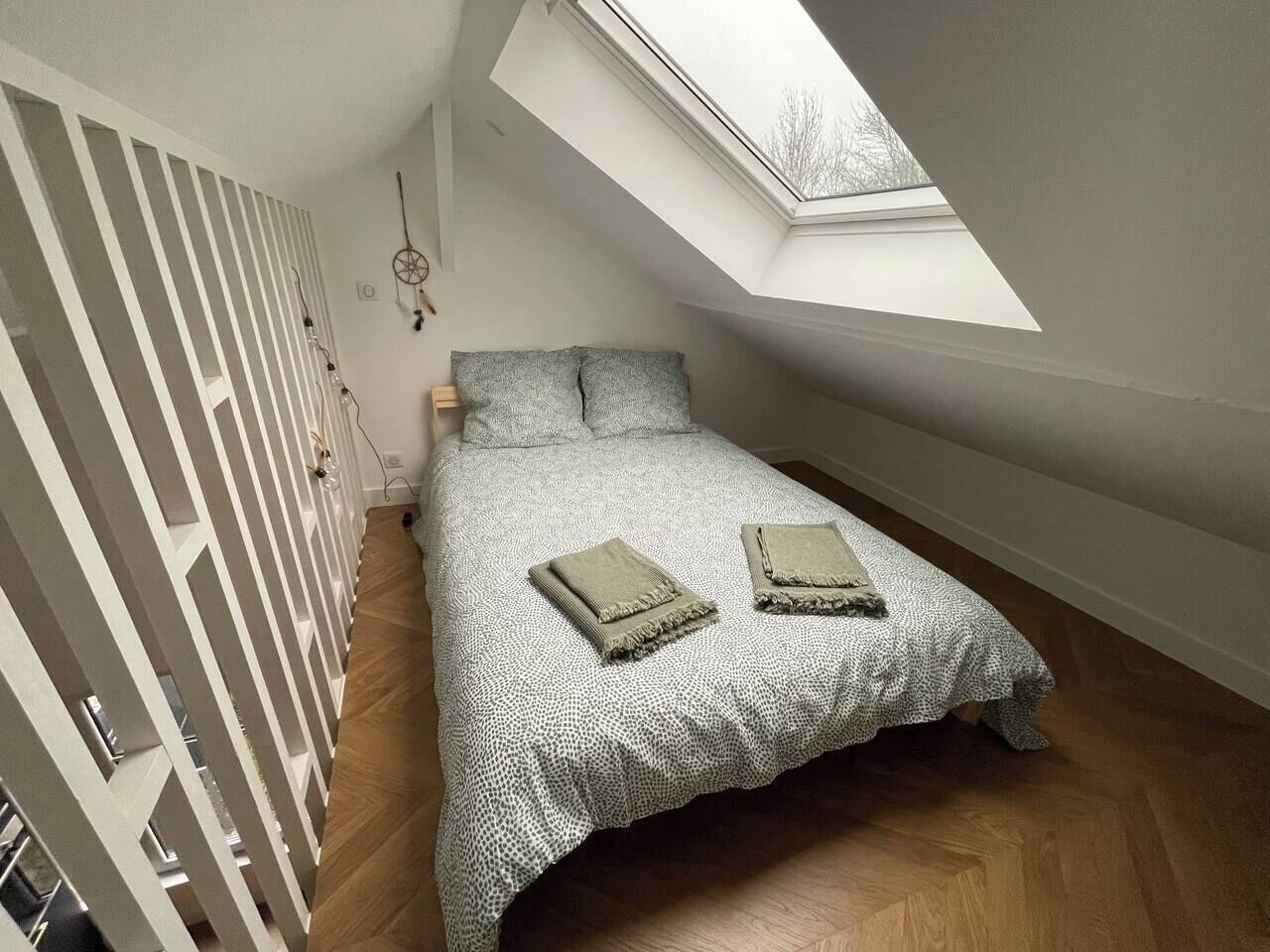 Ein kleines, gemütliches Schlafzimmer mit schrägem Dach und Dachfenster, mit einem ordentlich bezogenem Bett, zwei Kissen und zwei zusammengelegten Decken.