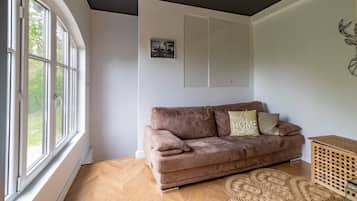 Een kamer met een groot raam, een bruine bank, een houten vloer en een aan de muur hangend schilderij.