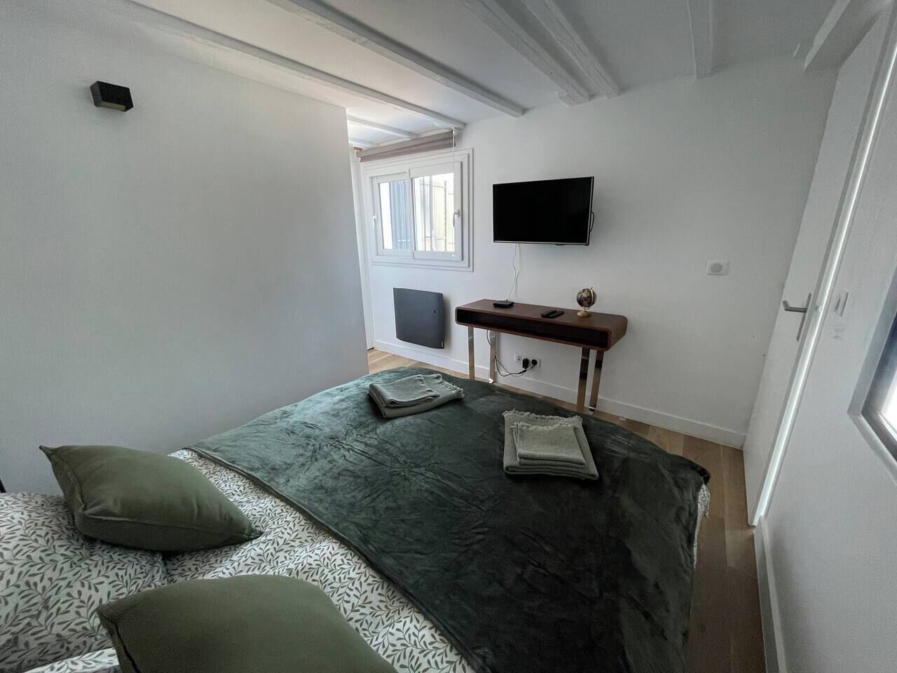 A bedroom with a bed, pillows, a desk, a chair, a flat-screen TV, and a window.