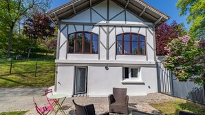 Une maison à deux étages, dotée d’un toit à pignon, d’un porche à l’avant et d’un petit patio aménagé avec du mobilier d’extérieur.