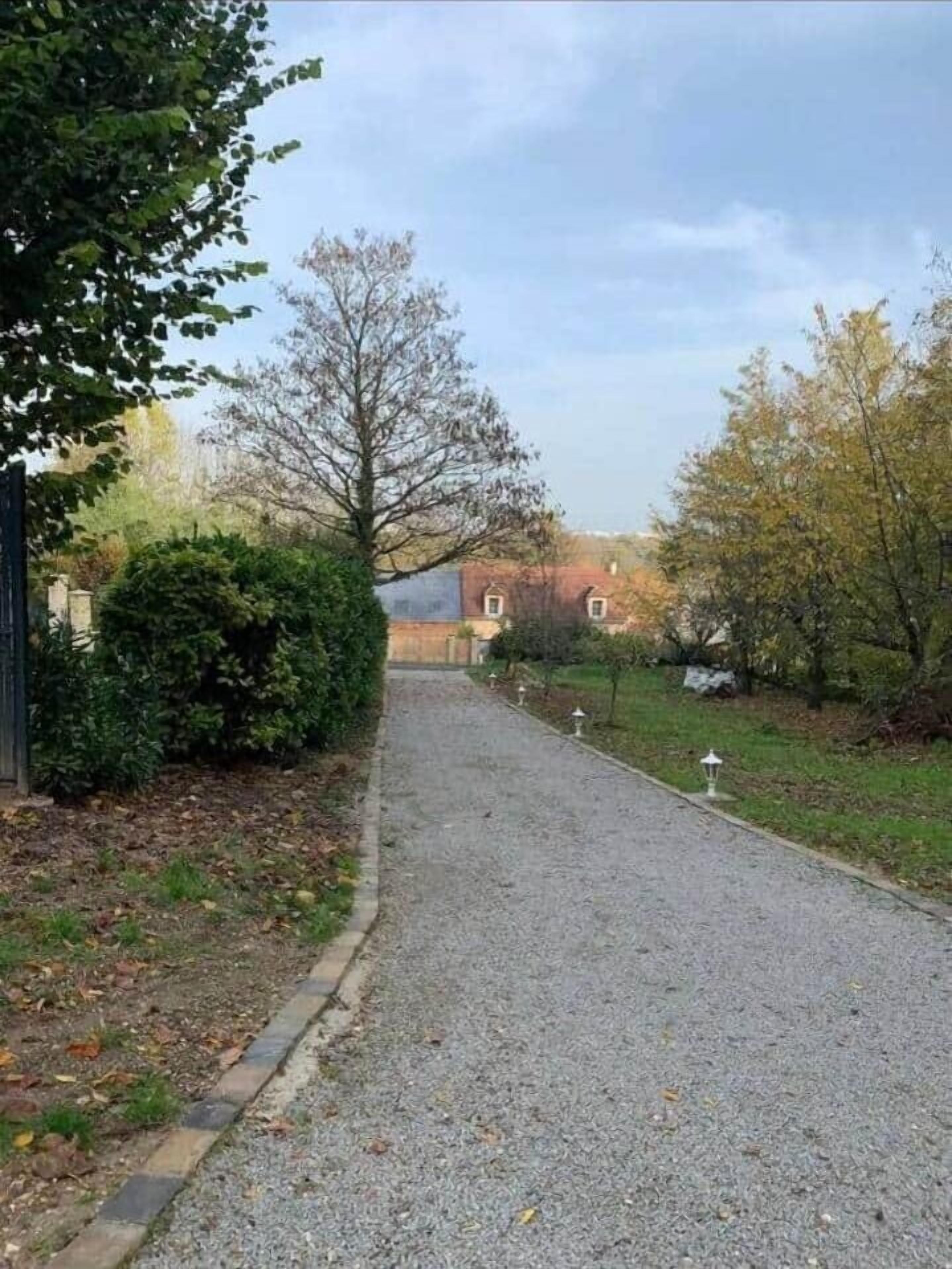 A gravel driveway leading to a building with a red roof, surrounded by trees and shrubs.