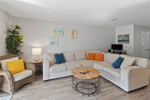 A living room with a sofa, a coffee table, a wicker chair, a lamp, and wall art. - Townhome · Walk to Beach · Screened Porch · Pools (Port St. Joe)