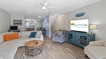 A modern living room with a sofa, armchair, coffee table, and a TV. There is a dining area with chairs and a table. The room has a ceiling fan and a sign on the wall.