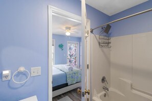 A bathroom with a shower, a bed with a blue and white floral bedspread, a ceiling fan, and a small window with a decorative tree. - Townhome · Walk to Beach · Screened Porch · Pools (Port St. Joe)