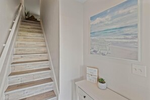 A staircase with wooden steps, a wall-mounted painting of beach chairs, and a sign that reads 'CATCH ME BY THE SEA.'