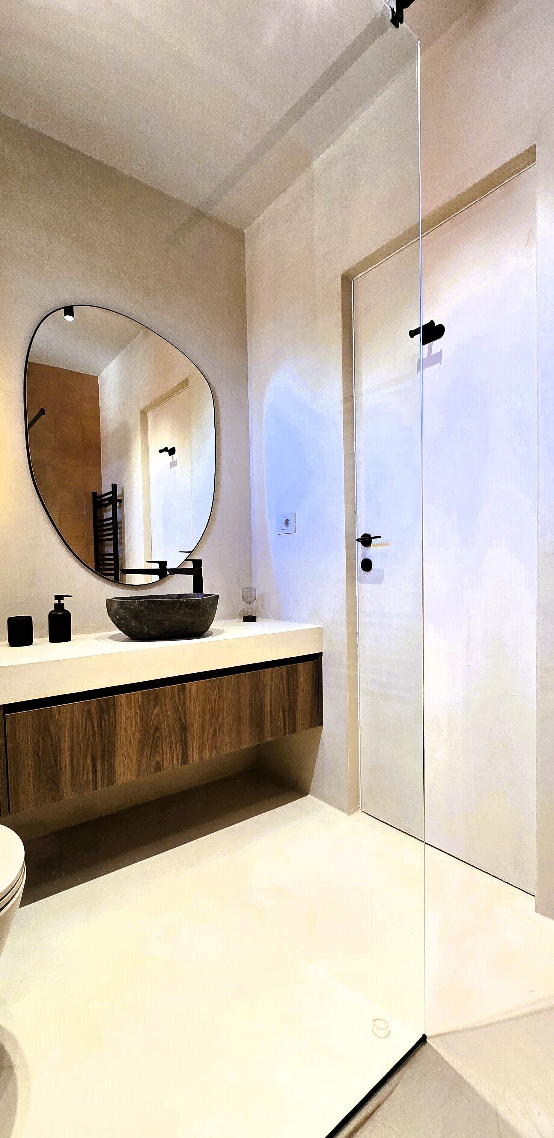 Un baño moderno con un espejo ovalado grande, un mueble de tocador de madera, un lavabo negro y una cabina de ducha con paredes de cristal.